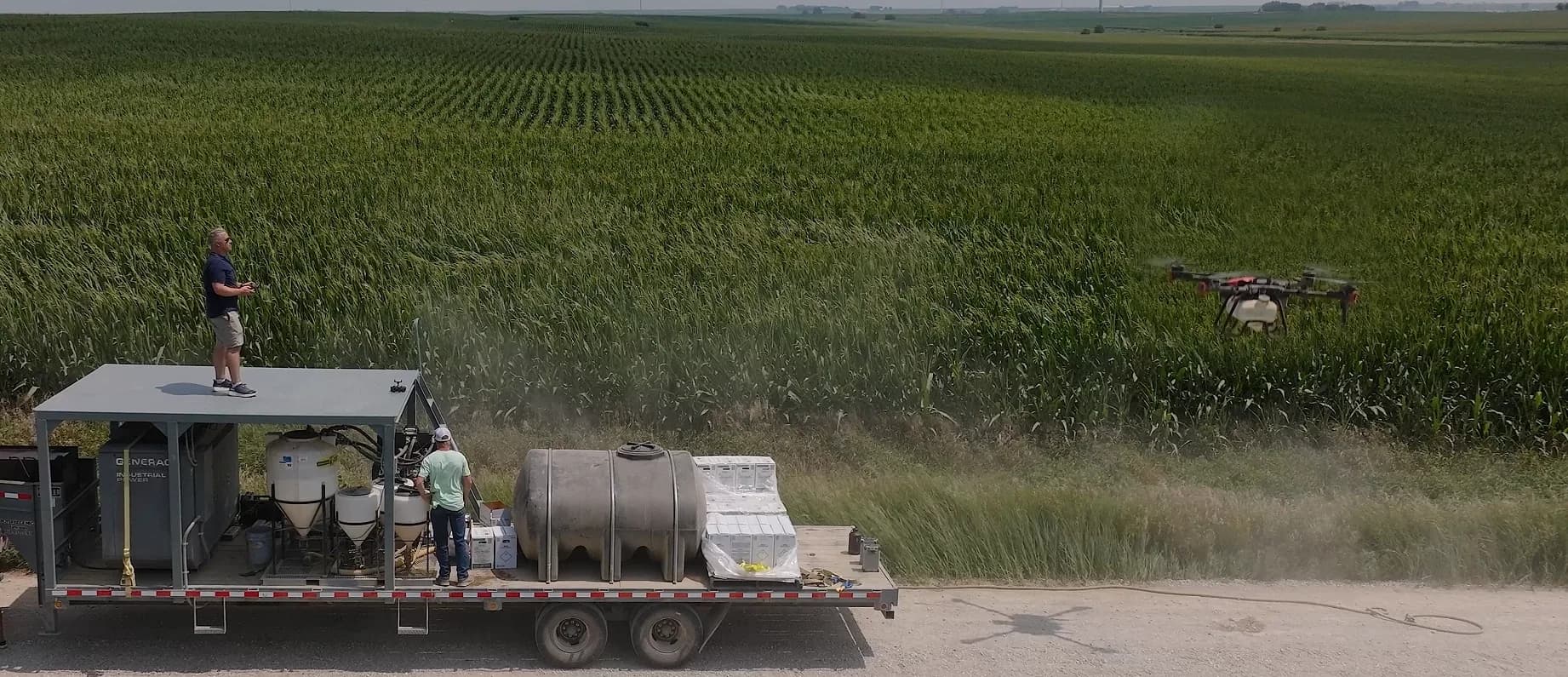 A Terraplex crew running a custom application from a trailer rig: a pilot flying from the deck while a spray drone works a cornfield